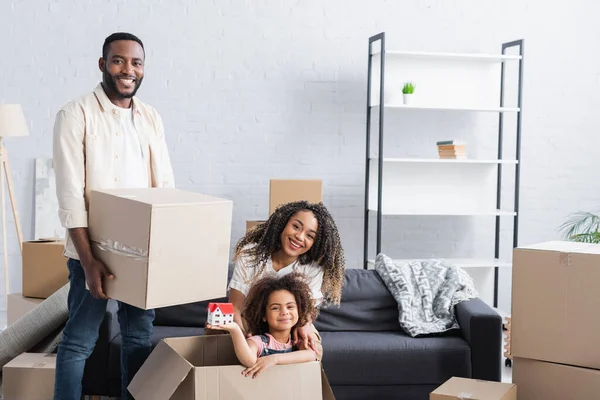 Chica afroamericana feliz en caja de cartón sosteniendo modelo de casa cerca de los padres en nuevo apartamento - foto de stock