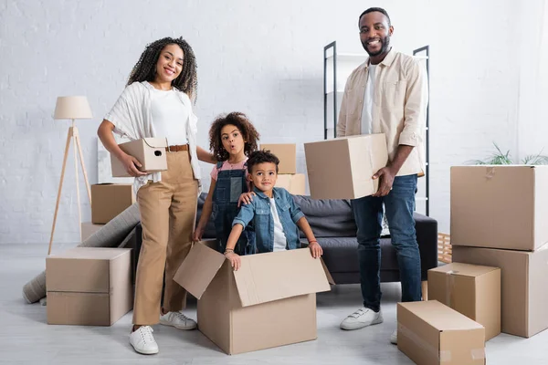 Frères et sœurs émerveillés dans une boîte en carton près des parents afro-américains avec des paquets — Photo de stock