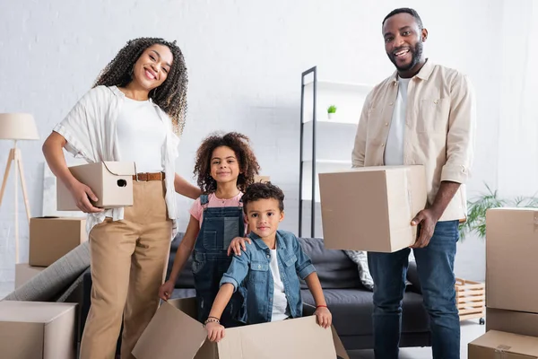 Alegres hermanos afroamericanos en caja de cartón cerca de padres con paquetes en nuevo apartamento - foto de stock