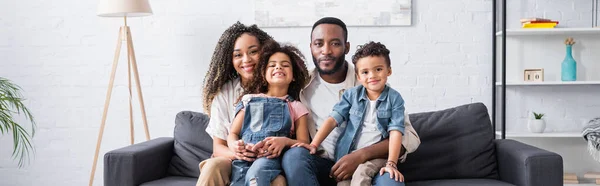 Gioiosa famiglia afroamericana sorridente alla macchina fotografica sul divano di casa, banner — Foto stock