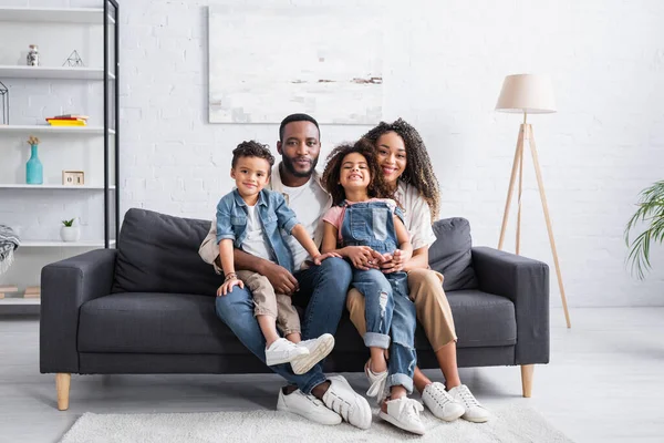 Familia afroamericana feliz sentado en el sofá en casa y mirando a la cámara - foto de stock