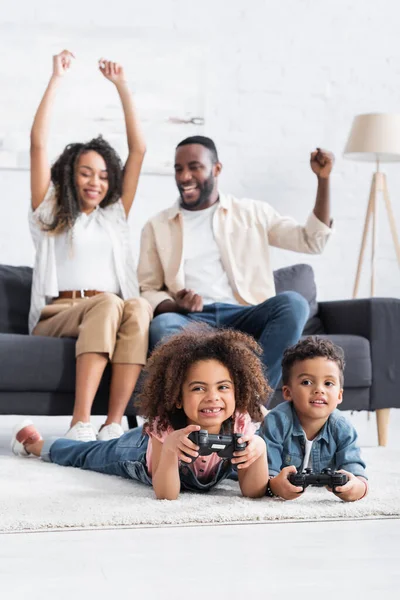 KYIV, UKRAINE - JANUARY 25, 2021: excited african american parents showing win gesture near kids gaming on floor — Stock Photo