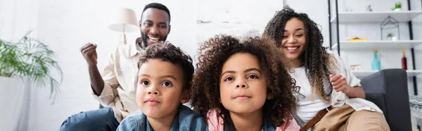 Hermanos afroamericanos concentrados viendo televisión cerca de padres alegres mostrando gesto de victoria, pancarta - foto de stock