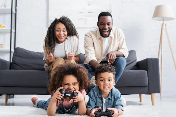KYIV, UCRANIA - 25 de enero de 2021: padres afroamericanos emocionados cerca de niños jugando videojuegos en el suelo - foto de stock