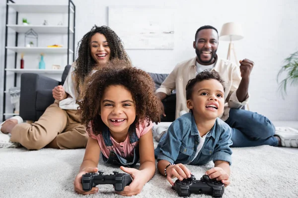 KYIV, UCRANIA - 25 de enero de 2021: niños afroamericanos emocionados jugando videojuegos en el suelo cerca de padres borrosos - foto de stock