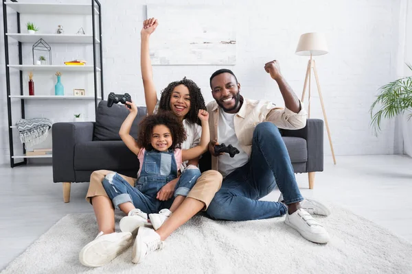 KYIV, UKRAINE - JANUARY 25, 2021: excited african american family showing triumph gesture while gaming on floor at home — Stock Photo
