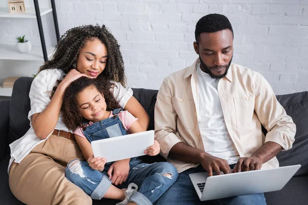 Африканский американец, печатающий на ноутбуке рядом с женой и дочерью с цифровым планшетом — стоковое фото