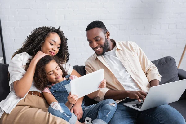 Uomo afroamericano sorridente digitando sul computer portatile e guardando la figlia con tablet digitale — Foto stock