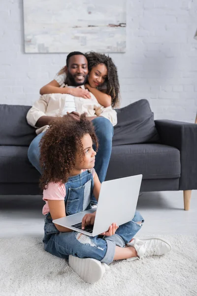 Happy african american woman embracing husband near daughter sitting on floor with laptop — Stock Photo