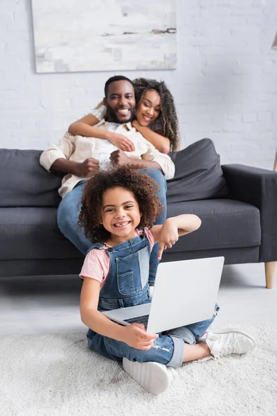 Glückliches afrikanisch-amerikanisches Mädchen zeigt auf Laptop, während es auf dem Boden in der Nähe verschwommener Eltern sitzt — Stockfoto