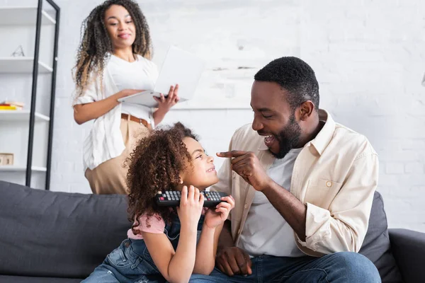 Cheerful african american man touching nose of daughter near blurred wife with laptop — Stock Photo
