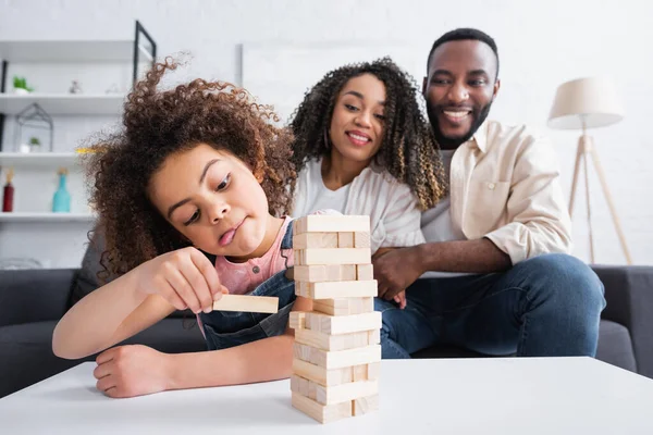 Africano americano chica sobresaliendo lengua mientras jugando madera bloques juego cerca borrosa padres - foto de stock