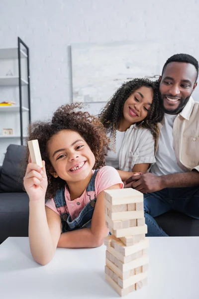 Joyeuse fille afro-américaine souriant à la caméra près de blocs de bois jeu et parents heureux — Photo de stock