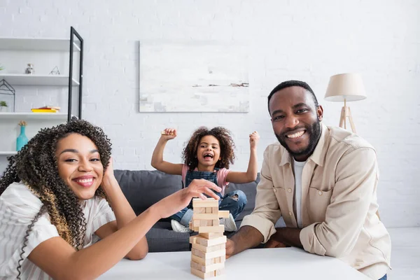 Animado afro-americano menina mostrando triunfo gesto perto sorrindo pai jogando madeira blocos jogo — Fotografia de Stock