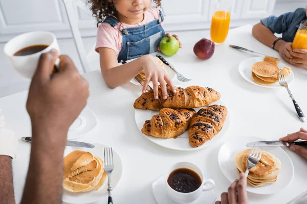 Abgeschnittene Ansicht eines afrikanisch-amerikanischen Mädchens, das beim Frühstück mit verschwommener Familie Croissant einnimmt — Stockfoto