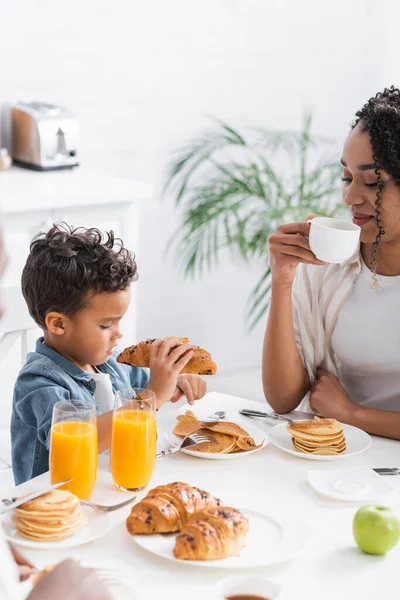 Africano americano chico holding croissant cerca mamá con taza de café - foto de stock