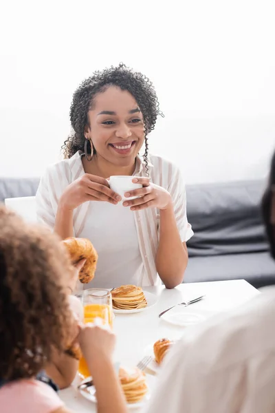 Sonriente afroamericana mujer sosteniendo taza de café durante el desayuno con la familia - foto de stock