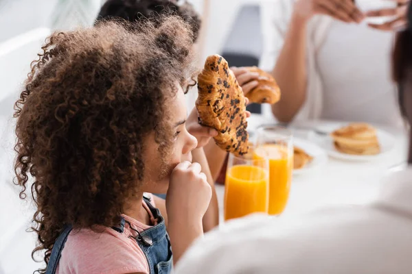 Menina americana africana segurando croissant perto de suco de laranja e família turva — Fotografia de Stock