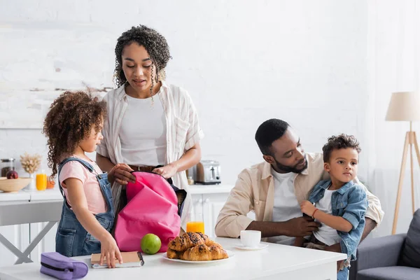 Mujer afroamericana embalaje mochila de hija en cocina - foto de stock