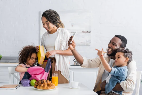 African american woman helping daughter packing backpack in kitchen — Stock Photo