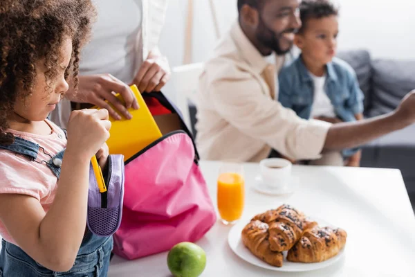 Woman packing backpack of african american daughter near blurred husband and son — Stock Photo