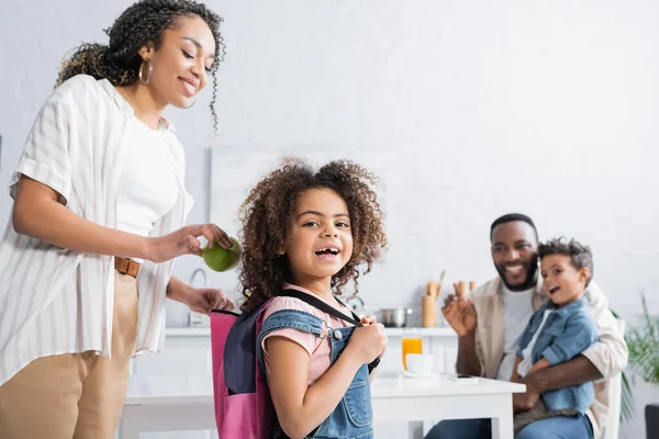 African american woman putting apple into backpack of daughter — Stock Photo