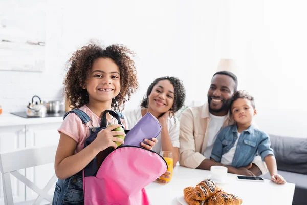 Menina americana africana alegre com caixa de lápis e mochila perto de família feliz na cozinha — Fotografia de Stock