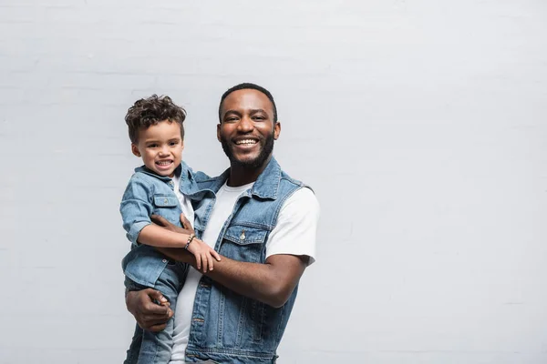 Happy african american man holding kid while looking at camera on grey — Stock Photo