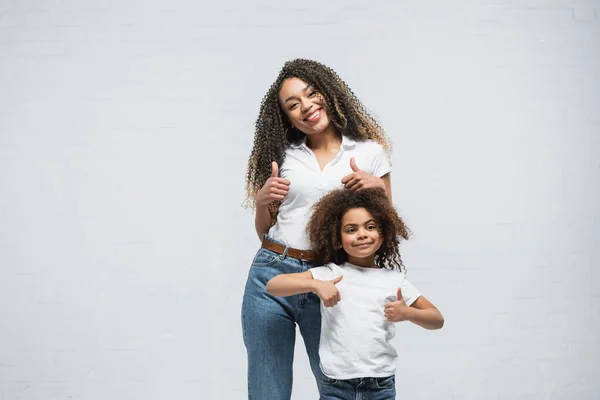 Mujer afroamericana positiva con el niño mostrando los pulgares hacia arriba en gris - foto de stock