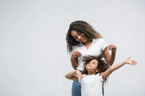 Freudige Frau berührt Haare der lockigen afrikanisch-amerikanischen Tochter auf grau — Stockfoto