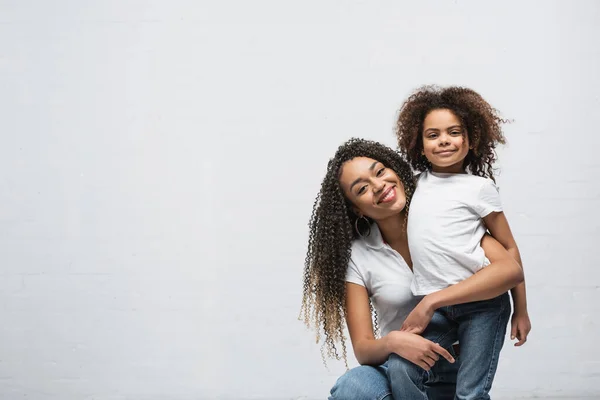 Happy african american woman embracing smiling daughter while looking at camera on grey — Stock Photo