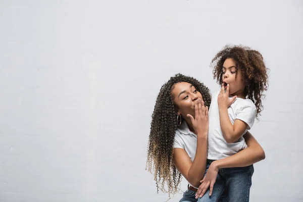 Erstaunt afrikanisch-amerikanische Mutter mit Kind, das Mund mit der Hand bedeckt und einander auf grau anschaut — Stockfoto