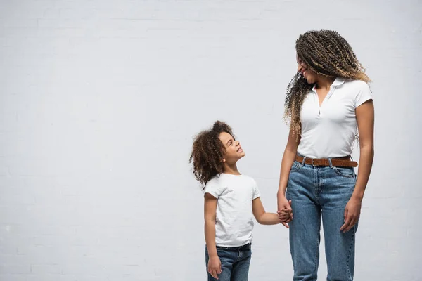 Happy african american woman with daughter looking at each other while holding hands on grey — Stock Photo