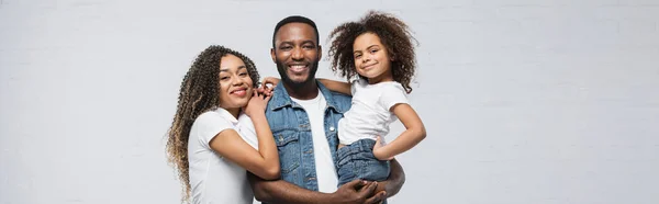 Uomo afroamericano sorridente che tiene il bambino vicino alla moglie felice sul grigio, striscione — Foto stock