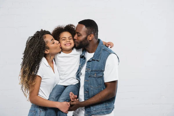 Glückliche afrikanisch-amerikanische Eltern küssen lächelnde Tochter auf grau — Stockfoto