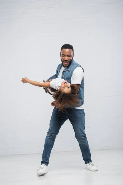 Cheerful african american man holding excited daughter in hands on grey — Stock Photo