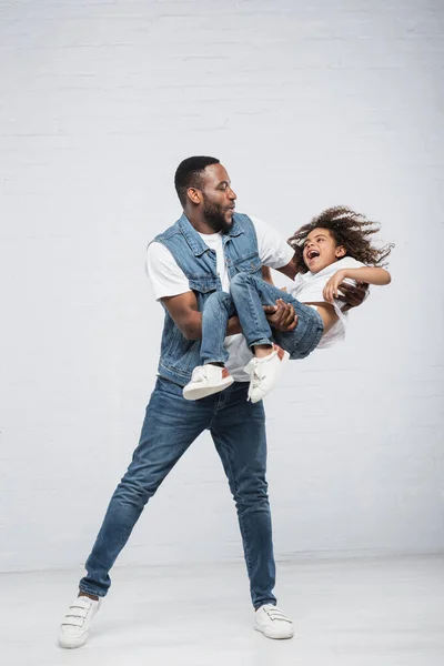 Happy african american man playing with thrilled daughter while holding her in hands on grey — Stock Photo