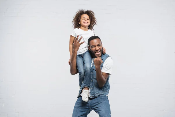 Überglücklicher afrikanisch-amerikanischer Mann mit glücklicher Tochter auf der Schulter, die Yah-Geste auf grau zeigt — Stockfoto