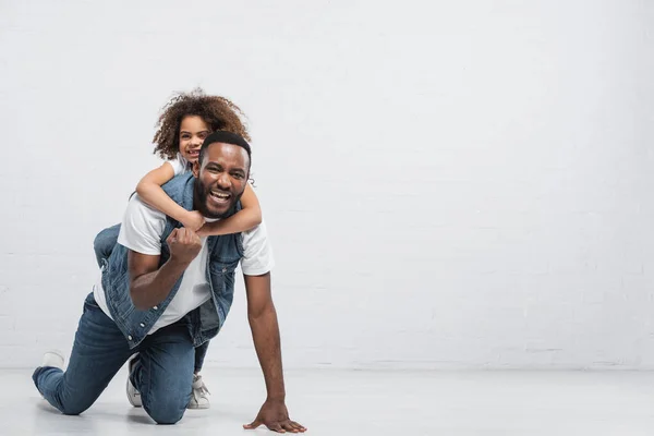 Cheerful african american girl piggybacking father showing win gesture while kneeling on grey — Stock Photo