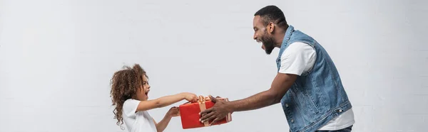 Vue de côté de l'homme afro-américain présentant un cadeau à la fille étonnée sur gris, bannière — Photo de stock