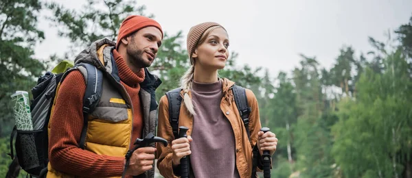 Coppia con zaini che reggono bastoni da trekking e distolgono lo sguardo nella foresta, banner — Foto stock