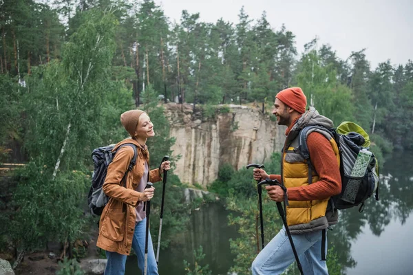 Счастливая пара с рюкзаками, держащими трости и смотрящими друг на друга возле озера — стоковое фото