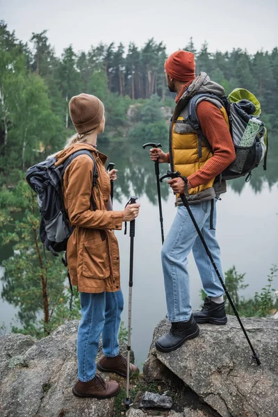 Vista trasera de pareja con mochilas sosteniendo bastones de senderismo y mirando al lago - foto de stock
