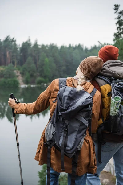Back view of woman with backpack holding hiking stick and leaning on man near at lake — Stock Photo