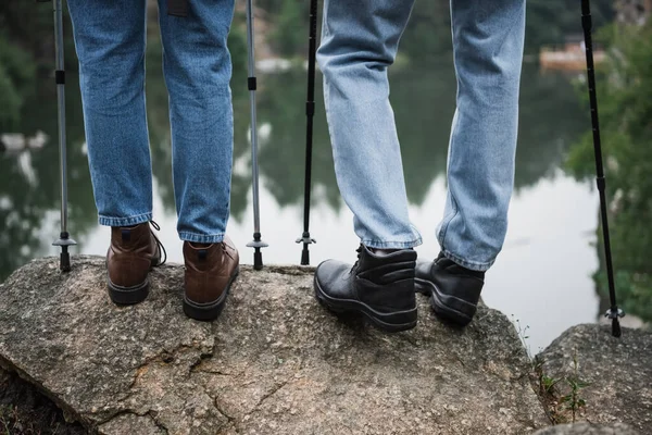 Vue partielle du jeune couple debout avec des bâtons de randonnée sur le rocher — Photo de stock