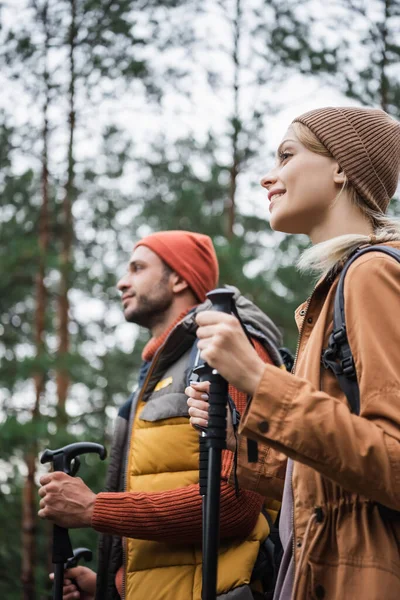 Alegre mujer en sombrero celebración senderismo palos cerca borrosa novio en el bosque - foto de stock