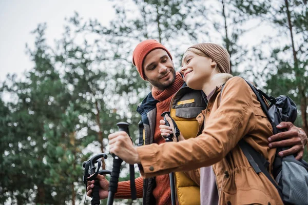 Happy man hugging girlfriend with hiking sticks in forest — Stock Photo