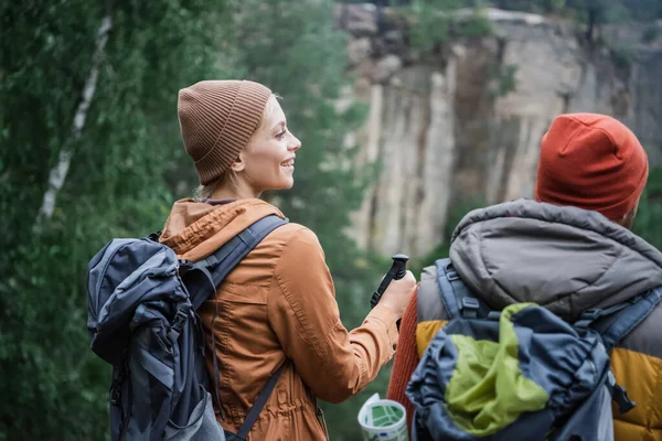 Cheerful young woman in hat standing with backpack near man in forest — Stock Photo