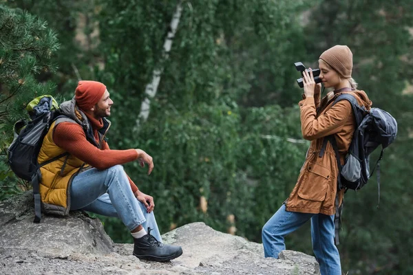 Mulher com mochila tirando foto de namorado feliz na câmera vintage na floresta — Fotografia de Stock