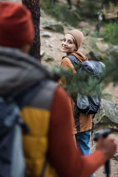 Happy woman with backpack looking at man trekking in forest on blurred foreground — Stock Photo